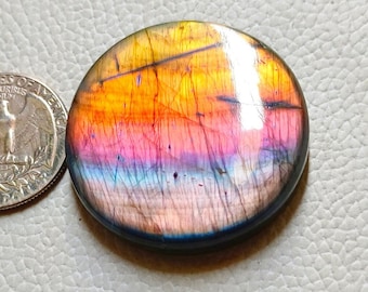 May include: A round, polished gemstone with a vibrant color gradient of yellow, orange, pink, and purple. A U.S. quarter coin is placed next to the gemstone for size comparison. The gemstone has a glossy finish and is set against a light gray surface.