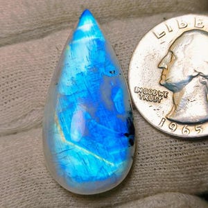 Op de afbeelding: Een druppelvormige, iriserende maansteen met blauwe en witte tinten. Een Amerikaanse kwartdollar uit 1965 ligt naast de steen ter vergelijking. De edelsteen ligt tegen een getextureerde, gebroken witte achtergrond.