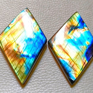 May include: Two diamond-shaped labradorite gemstones. The stones display a vibrant play of color, with hues of blue, yellow, and orange. The gemstones are polished and have a glossy finish, showcasing their natural beauty. The stones are side by side.