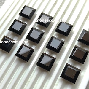 May include: A selection of square-cut, black gemstones displayed on a white, ridged surface. The gemstones are highly polished, reflecting light, and are evenly spaced. The text "stonescompany" is visible on the surface.