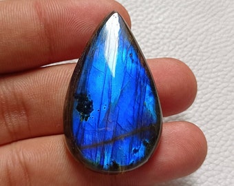 May include: A teardrop-shaped labradorite gemstone with a polished surface. The stone displays a vibrant blue iridescence with streaks of darker blue and black. The gemstone is held in a hand.