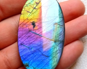 May include: An oval-shaped labradorite gemstone with a vibrant display of colors. The stone features a mix of purple, blue, green, and yellow hues, with a shimmering, iridescent effect. The gemstone is held in a hand, showcasing its polished surface and natural patterns.
