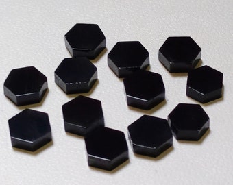 Può includere: Una collezione di dodici gemme esagonali nere. Ogni pietra ha una superficie lucida e riflettente. Le gemme sono disposte su una superficie chiara, mettendo in risalto il loro colore scuro e la forma geometrica.