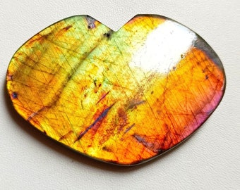 May include: A heart-shaped labradorite gemstone with a vibrant display of colors. The stone features a mix of yellow, orange, purple, and green hues, with a polished surface. A US quarter is next to the stone for size comparison.
