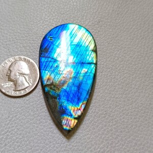 May include: A teardrop-shaped labradorite gemstone with vibrant blue, green, and yellow iridescence. The polished stone is next to a quarter for size comparison, showcasing its elongated form and natural patterns. The gemstone is set against a gray surface.