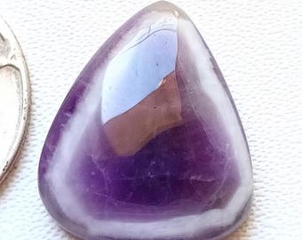 May include: A polished, teardrop-shaped gemstone with a deep purple hue and white banding. The stone is next to a U.S. quarter for size comparison. The gemstone has a smooth, reflective surface.