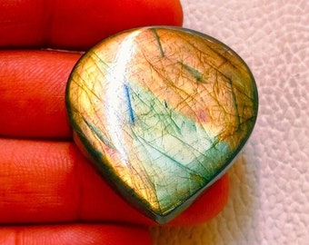 May include: A polished, teardrop-shaped gemstone with a mix of colors, including gold, green, and blue. The stone has a smooth surface and a natural, iridescent appearance. The gemstone is held in a hand.