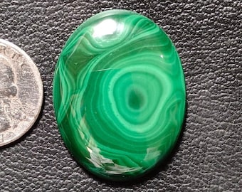 May include: Oval-shaped polished malachite gemstone with swirling green patterns. A U.S. quarter coin is next to the gemstone for size comparison. The stone is on a black textured surface.