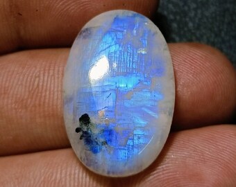 Può includere: Pietra di luna ovale, con una base bianca perlacea e iridescenza blu interna. La superficie lucida mostra riflessi blu, con alcune inclusioni scure. La pietra è tenuta in una mano.