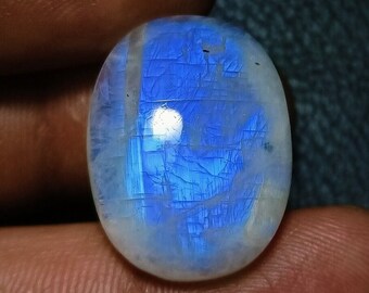 Può includere: Pietra di luna ovale con una lucentezza iridescente blu e bianca. La superficie lucida presenta fratture interne, creando un motivo unico. La gemma è tenuta tra le dita su uno sfondo scuro.