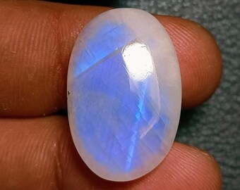 Può includere: Pietra preziosa a forma ovale di pietra di luna con superficie lucida. La pietra presenta una base bianco latte con riflessi blu iridescenti. La gemma è tenuta in mano, mostrando la sua bellezza naturale e il suo potenziale per la gioielleria.