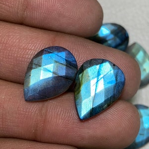 Puede incluir: Dos piedras preciosas de labradorita facetadas en forma de lágrima. Las piedras muestran un juego de colores con azules, verdes y púrpuras. Las gemas se sostienen en una mano, con otras piedras similares visibles en el fondo.