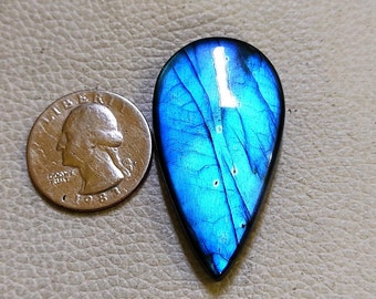 May include: A teardrop-shaped, polished labradorite gemstone with vibrant blue iridescence. The stone is next to a U.S. quarter for size comparison. The gemstone's surface displays a play of color, with streaks of blue and hints of other hues.