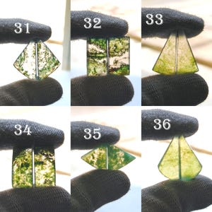 May include: A collection of six moss agate gemstone cabochons in various shapes: diamond, rectangular, and triangular. The gemstones feature green and white patterns, held by a gloved hand. Each stone is numbered from 31 to 36.
