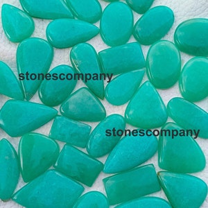 May include: A collection of polished, teal-colored gemstones in various shapes, including ovals, rectangles, and teardrops. The gemstones are scattered across a light-colored surface, with the text "stonescompany" visible on some.