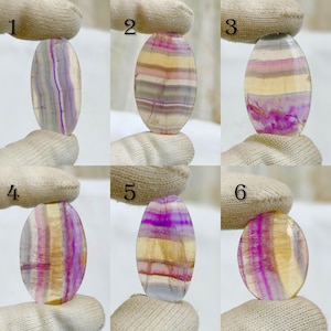 Può includere: Una collezione di sei pietre preziose di fluorite di forma ovale. Ogni pietra mostra un motivo di colori unico, tra cui viola, giallo e bianco. Le pietre sono presentate su uno sfondo neutro, evidenziando la loro bellezza naturale e le variazioni.