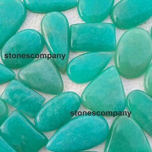 May include: A collection of polished, turquoise-colored gemstones in various shapes, including ovals, rectangles, and teardrops. The gemstones have a smooth, glossy surface. The text "stonescompany" is visible on some of the stones.