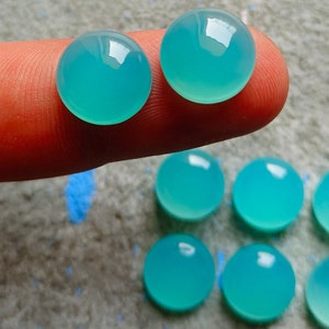 K&ouml;nnte beinhalten: Eine Sammlung runder, polierter, aquafarbener Edelsteine. Zwei Edelsteine werden auf einem Finger gehalten, mit mehreren weiteren darunter angeordnet. Die Steine haben eine glatte, reflektierende Oberfl&auml;che und die Farbe ist ein lebendiges, helles T&uuml;rkis.