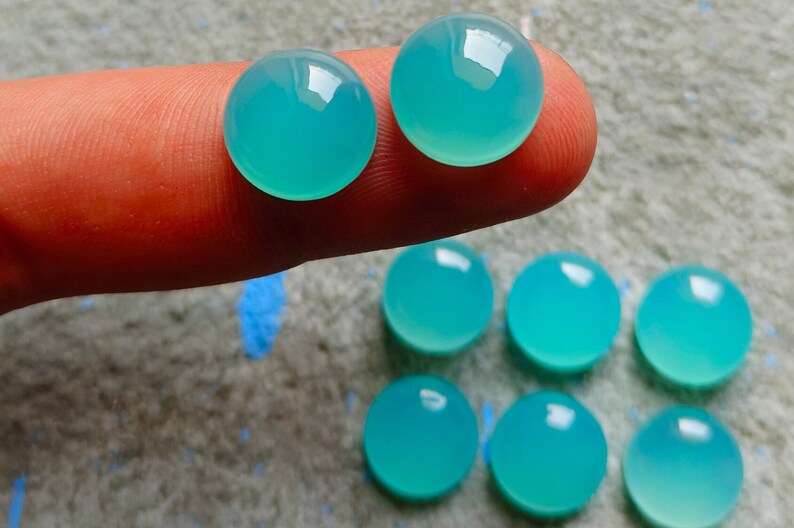 K&ouml;nnte beinhalten: Eine Sammlung runder, polierter, aquafarbener Edelsteine. Zwei Edelsteine werden auf einem Finger gehalten, mit mehreren weiteren darunter angeordnet. Die Steine haben eine glatte, reflektierende Oberfl&auml;che und die Farbe ist ein lebendiges, helles T&uuml;rkis.