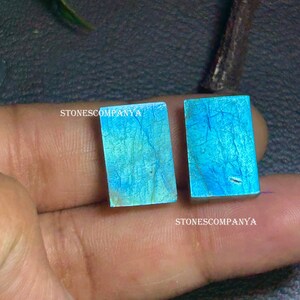 Pode incluir: Duas pedras preciosas retangulares polidas, com uma cor azul-esverdeada vibrante e veios intrincados. As pedras são seguradas em uma mão, com o texto "STONESCOMPANYA" visível. Estas pedras são provavelmente usadas para joias ou fins decorativos.