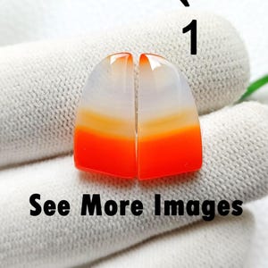 以下が含まれることがあります： 磨かれたアゲート宝石カボション2つ。各カボションは半月形で、上部が白、オレンジに変わり、鮮やかな赤で終わる色のグラデーションがあります。「See More Images」の文字が下にあります。
