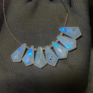 May include: A necklace featuring seven faceted, iridescent moonstone pendants. The pendants are a light blue and white color, with flashes of blue. Small, gold-colored beads separate the moonstone pendants. The necklace is strung on a thin, silver-colored wire.