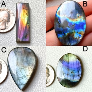 May include: Four polished labradorite gemstones in various shapes: rectangular, oval, and teardrop. The stones display iridescent flashes of blue, purple, and yellow. A quarter coin is next to each stone for size comparison. The gemstones are on a light-colored surface.