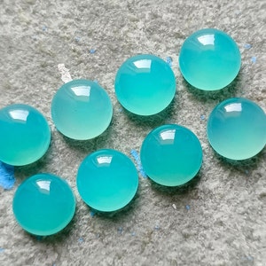 K&ouml;nnte beinhalten: Acht runde, polierte, aquamarinfarbene Edelsteine liegen auf einer strukturierten, grauen Oberfl&auml;che. Die Edelsteine haben eine transluzente Qualit&auml;t und reflektieren das Licht. Das Arrangement ist minimalistisch und betont die nat&uuml;rliche Sch&ouml;nheit der Steine.