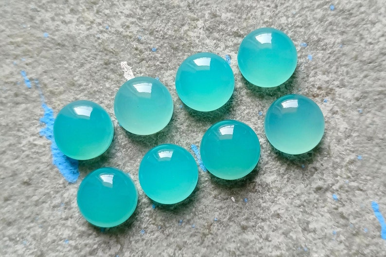 K&ouml;nnte beinhalten: Acht runde, polierte, aquamarinfarbene Edelsteine liegen auf einer strukturierten, grauen Oberfl&auml;che. Die Edelsteine haben eine transluzente Qualit&auml;t und reflektieren das Licht. Das Arrangement ist minimalistisch und betont die nat&uuml;rliche Sch&ouml;nheit der Steine.