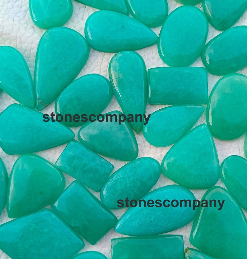 May include: A collection of polished, turquoise-colored gemstones in various shapes, including teardrops, rectangles, and ovals. The gemstones are scattered across a white surface, with the text "stonescompany" visible on some of them.