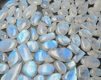 May include: A collection of polished moonstone gemstones in various shapes, including oval, teardrop, and rectangular. The stones display a milky white base with iridescent blue flashes. The gemstones are arranged closely together, filling the frame.