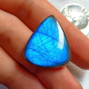 May include: A triangular, polished labradorite gemstone with a vibrant blue hue and internal striations. The stone is held in a hand, showcasing its iridescent quality. A silver coin is visible in the background.