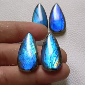 May include: Four teardrop-shaped labradorite gemstones. The stones display a vibrant play of color, with blues and hints of yellow and green. The polished surfaces catch the light, showcasing the natural patterns and iridescence of the gemstones.
