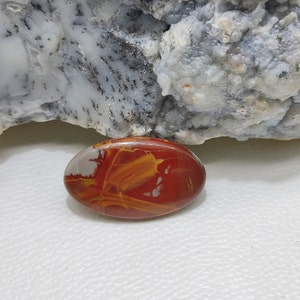 May include: Oval-shaped polished stone with red, yellow, and white hues. The stone has a smooth, glossy surface. It is set against a backdrop of a textured, light-coloured rock formation.