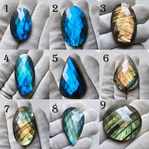 Op de afbeelding: Een verzameling van negen gefacetteerde labradoriet edelstenen in verschillende vormen. De stenen vertonen een scala aan kleuren, waaronder blauw, groen en goud, met een glinsterend, iriserend effect. Elke steen wordt in een gehandschoende hand gehouden.