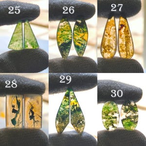 May include: A collection of six pairs of polished gemstone cabochons, each held by a gloved hand. The stones display a variety of colors, including green, yellow, and brown, with unique patterns and shapes. The cabochons are numbered 25 through 30.