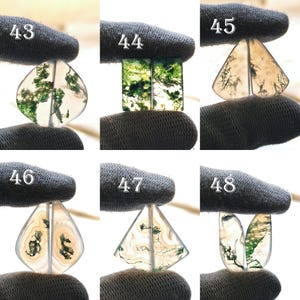 May include: A collection of six polished moss agate gemstone pairs, each with unique patterns of green moss-like inclusions. The stones are held by a gloved hand, showcasing their translucent quality and varied shapes, including teardrop, rectangular, and triangular forms.