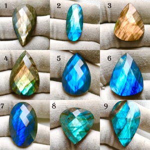 Op de afbeelding: Negen gefacetteerde labradoriet edelstenen in verschillende vormen en kleuren. De edelstenen variëren van druppelvormig tot ovaal en driehoekig, met kleuren als blauw, groen, goud en bruin. Elke steen heeft een uniek patroon en slijpsel.