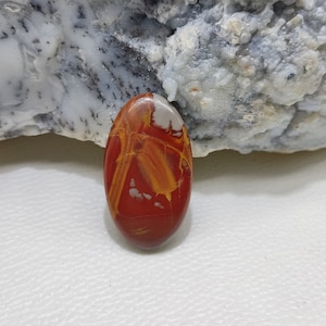 May include: Oval-shaped polished gemstone with a striking pattern of red, yellow, and white hues. The stone's surface displays a network of veins and colour variations. The gemstone is set against a neutral background, highlighting its natural beauty.