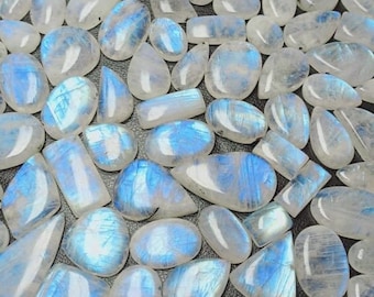 May include: A collection of polished moonstone gemstones in various shapes, including oval, teardrop, and rectangular. The stones have a translucent, milky white base with blue and iridescent flashes. The gemstones are scattered on a dark surface.