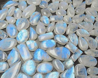 Puede incluir: Una colección de piedras lunares pulidas en varias formas, incluyendo ovaladas, en forma de lágrima y rectangulares. Las piedras son translúcidas con una base blanca lechosa y exhiben adularescencia azul. Las piedras preciosas están dispersas sobre una superficie oscura.