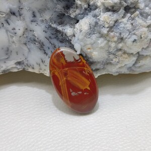 May include: Oval-shaped polished gemstone in red, yellow, and white. The stone has a smooth, glossy surface with a unique pattern of veins and swirls. The gemstone is likely a decorative item or for jewellery. The stone is approximately 4 cm in length.