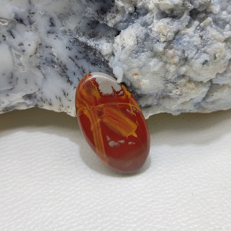 May include: Oval-shaped polished gemstone in red, yellow, and white. The stone has a smooth, glossy surface with a unique pattern of veins and swirls. The gemstone is likely a decorative item or for jewellery. The stone is approximately 4 cm in length.