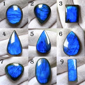 May include: A collection of nine polished labradorite gemstones in various shapes: oval, teardrop, rectangular, and more. The stones display a vibrant blue iridescence with internal striations. Each stone is unique, showcasing natural variations in color and pattern.