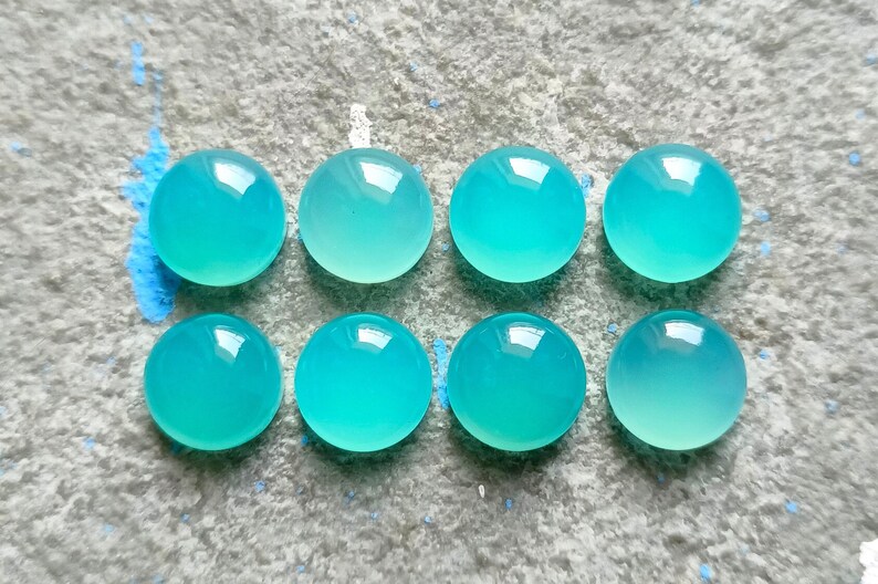 K&ouml;nnte beinhalten: Acht runde, polierte Cabochons in einem lebendigen, durchscheinenden T&uuml;rkis sind auf einer strukturierten, hellgrauen Oberfl&auml;che angeordnet. Die Edelsteine haben eine glatte, gl&auml;nzende Oberfl&auml;che, die das Licht reflektiert. Die Anordnung deutet auf eine Sammlung von Edelsteinen hin.
