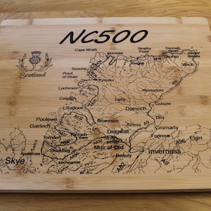 Può includere: Un tagliere in legno con una mappa incisa del percorso NC500 in Scozia. La mappa include i nomi delle città e dei paesi lungo il percorso, nonché i numeri delle strade principali.