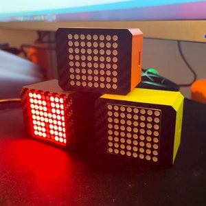 May include: Three small, square-shaped devices with black frames and colorful dot matrix displays. The top device is orange, the middle device is red, and the bottom device is yellow.