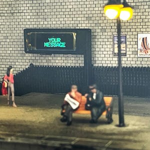 May include: A black and white image of a miniature city scene with a sign that reads "YOUR MESSAGE" in green text. Two figures are sitting on a bench in front of the sign.