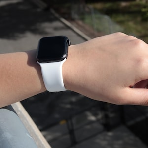 Op de afbeelding: Een zwarte smartwatch met een witte siliconen band wordt om een pols gedragen. Het horloge heeft een rechthoekig scherm en een zwarte rand. De witte band is glad en flexibel. Het horloge wordt buiten in natuurlijk licht getoond.