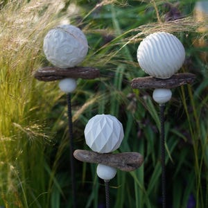 May include: Three garden ornaments with white spherical designs. Each stake has a different textured sphere: one with swirls, one with ridges, and one with a geometric pattern. The spheres are mounted on driftwood and supported by a white ball.